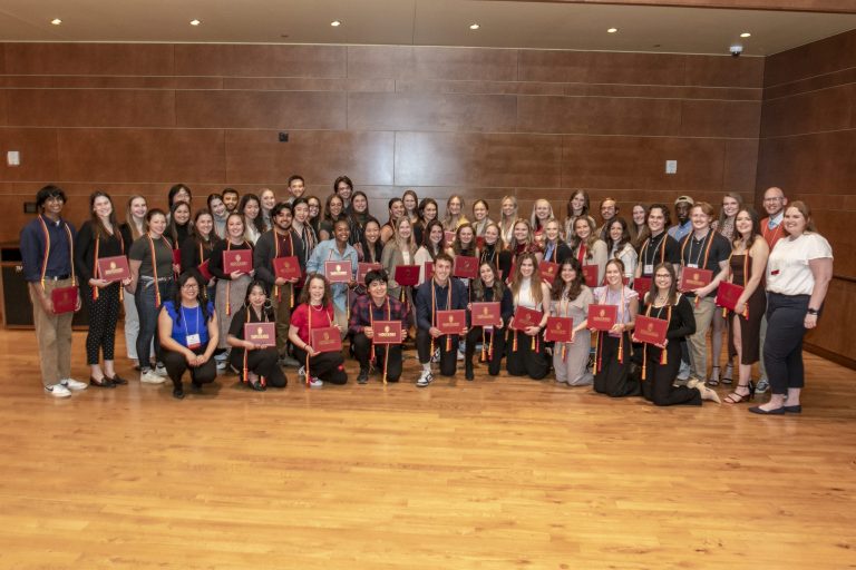 Group of 100 college students holding red certificate holders and wearing red and gold honor cords.