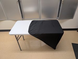 A four-foot table with a black table lay half covering it to show what the table lay looks like.