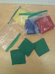 Three green bean bags outside of a ziplock bag with three other sets of three bean bags in red, blue, and yellow in the background.
