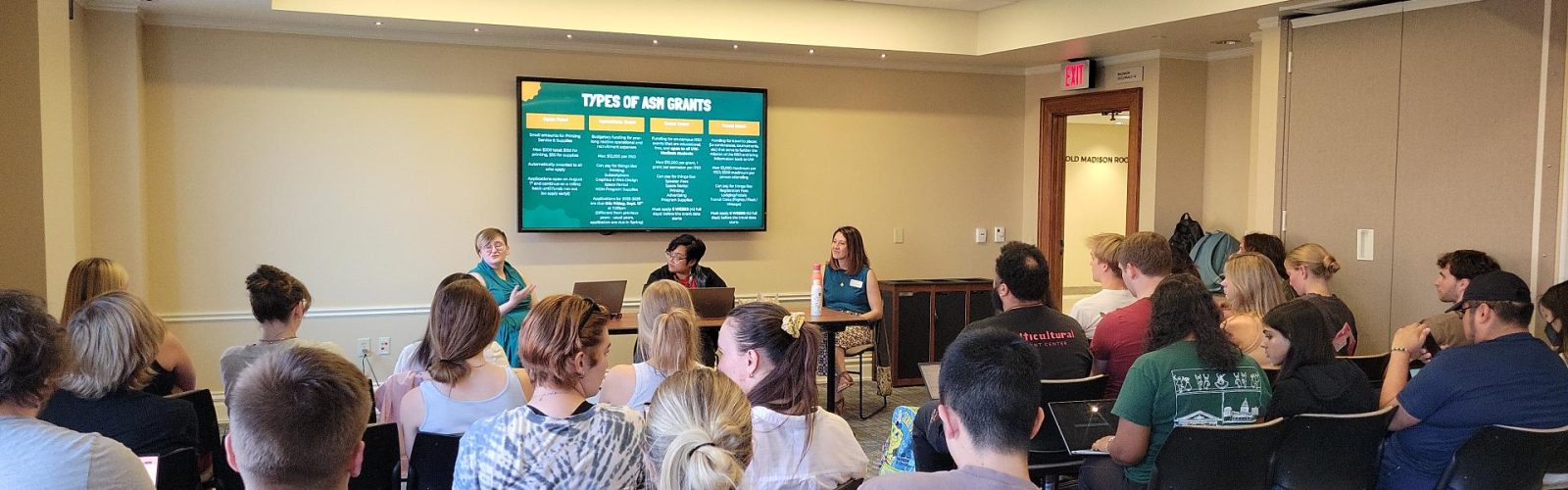 A room full of students listen to a presentation on grant opportunities for registered student organizations. Three of the grant representatives sit at the front with a teal-and-yellow themed PowerPoint slide behind them.