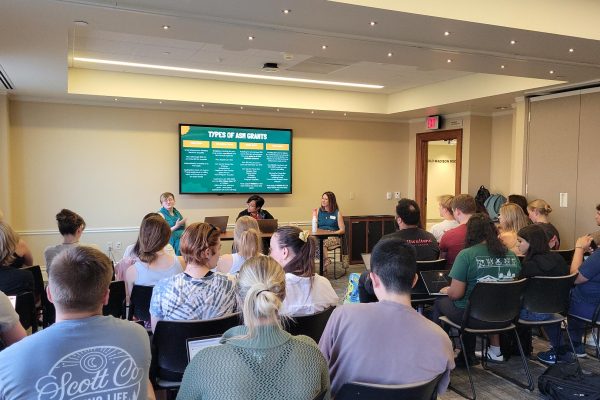 A room full of students listen to a presentation on grant opportunities for registered student organizations. Three of the grant representatives sit at the front with a teal-and-yellow themed PowerPoint slide behind them.