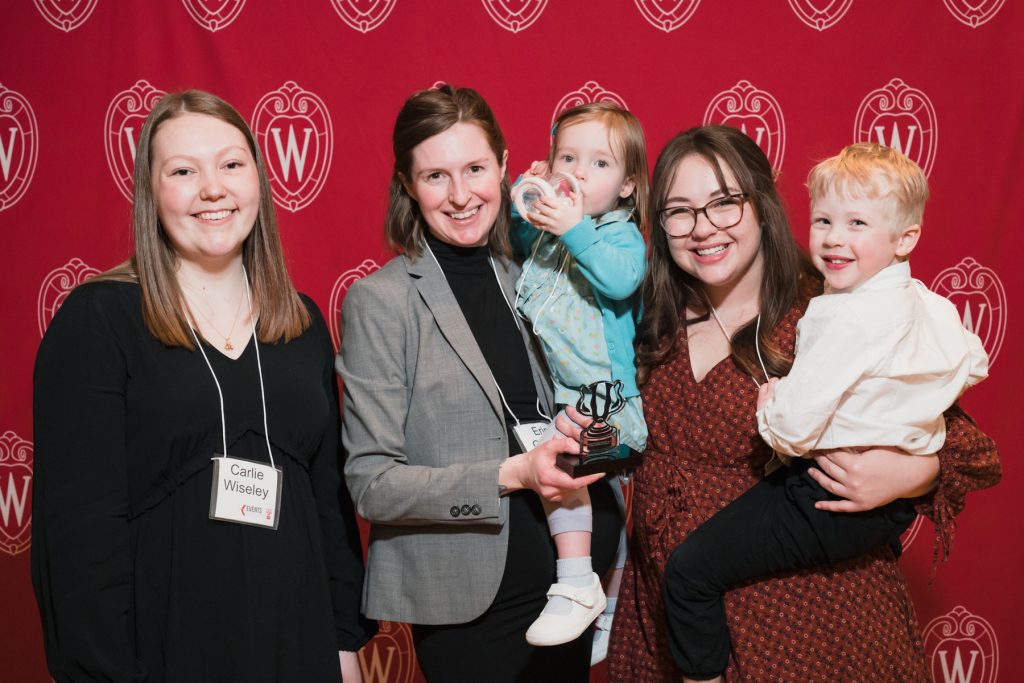 Wisconsin Student Parents Organization members pose holding their Bucky trophy on a red UW logo background. One woman is wearing a black top, another wearing a black blouse and grey jacketing, holding a toddler wearing a blue outfit drinking out of a sippy cup and another woman wearing a dark red dress holding a toddler wearing a white jacket. Everyone is smiling.
