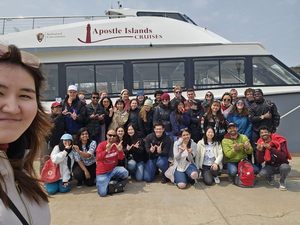 A selfie of a group of 35 students wearing warm outerwear pose in front of a white tour boat with dark-red text that reads 