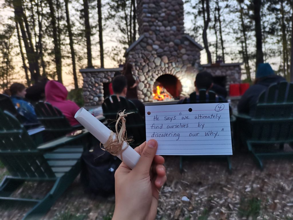 A participant holds an index card with a quote from the day in front of an outdoor fireplace by the tree line. It reads: He says 