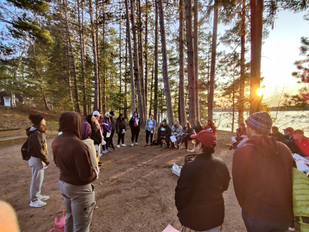 30 students stand in a circle by tree line and lakeside during sunset in a large-group discussion and debrief.