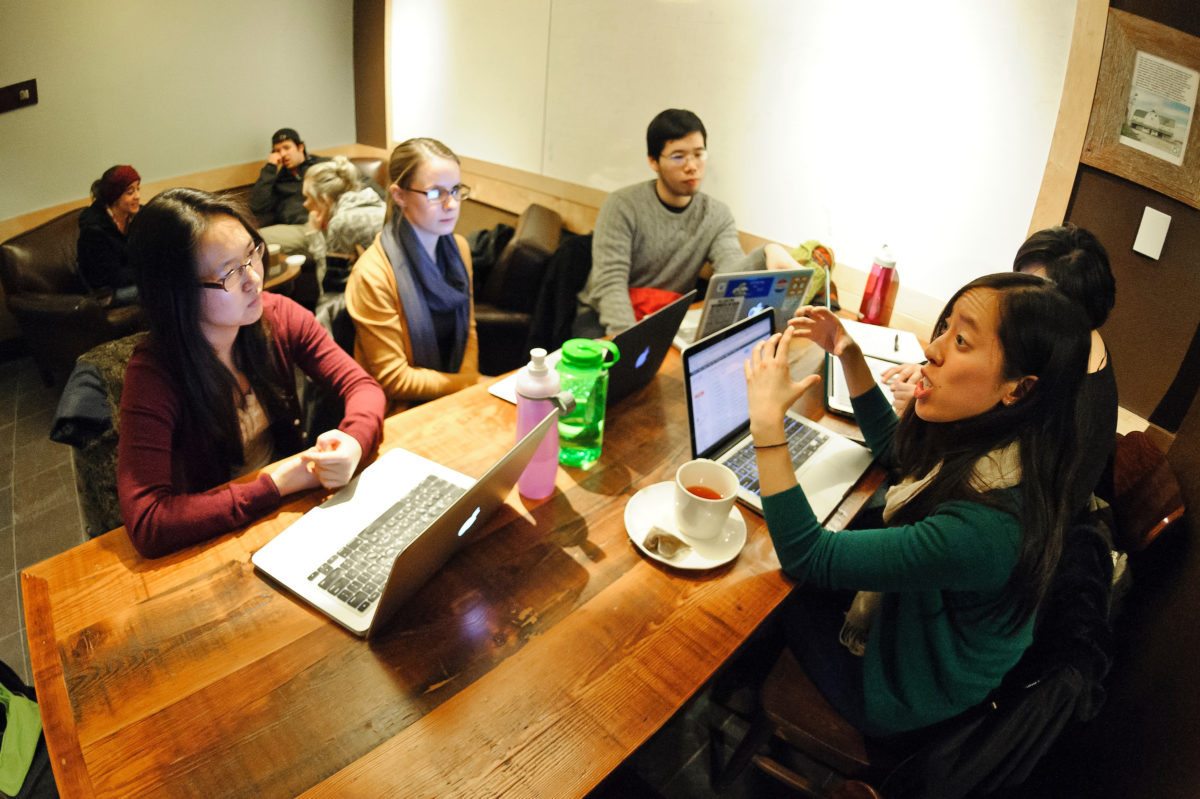 Group of 5 students sitting at a table with laptops in a discussion.