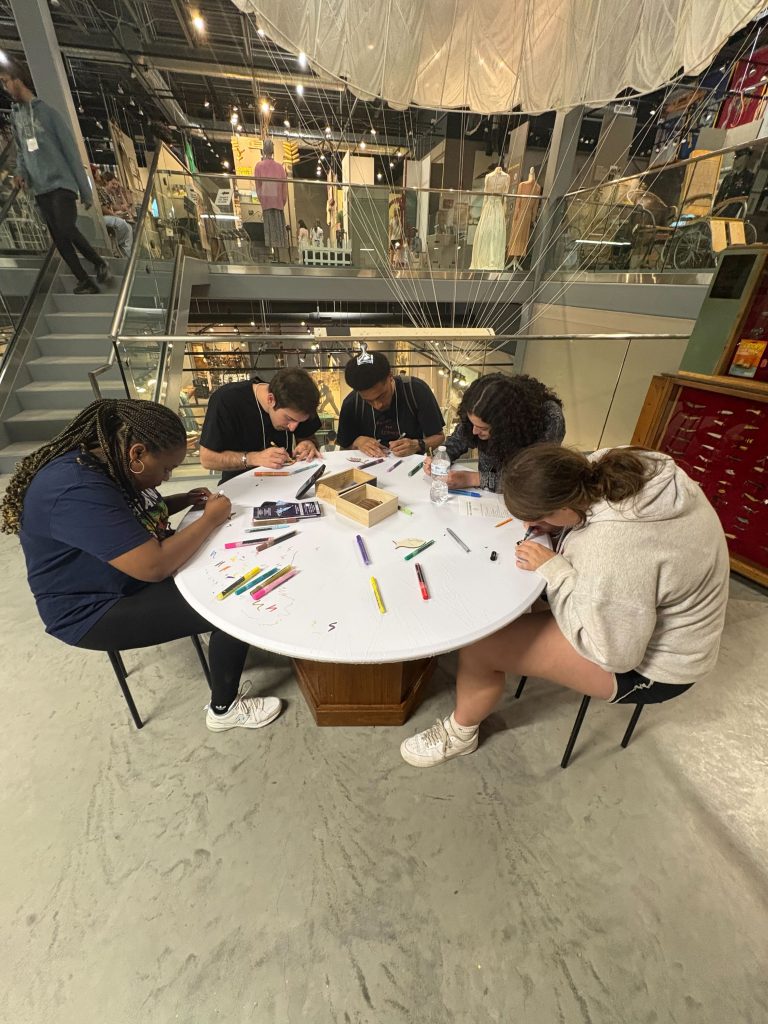 A group of five 2025 WEBT students sit at a circular, white table and design their own wooden fish trinkets during a visit to the Chippewa Area History Museum.