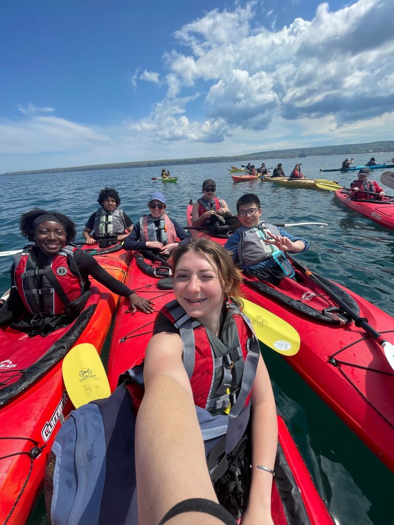Student Coordinator Fiona and 2025 WEBT students smiling in red kayaks on Lake Superior on a sunny day.