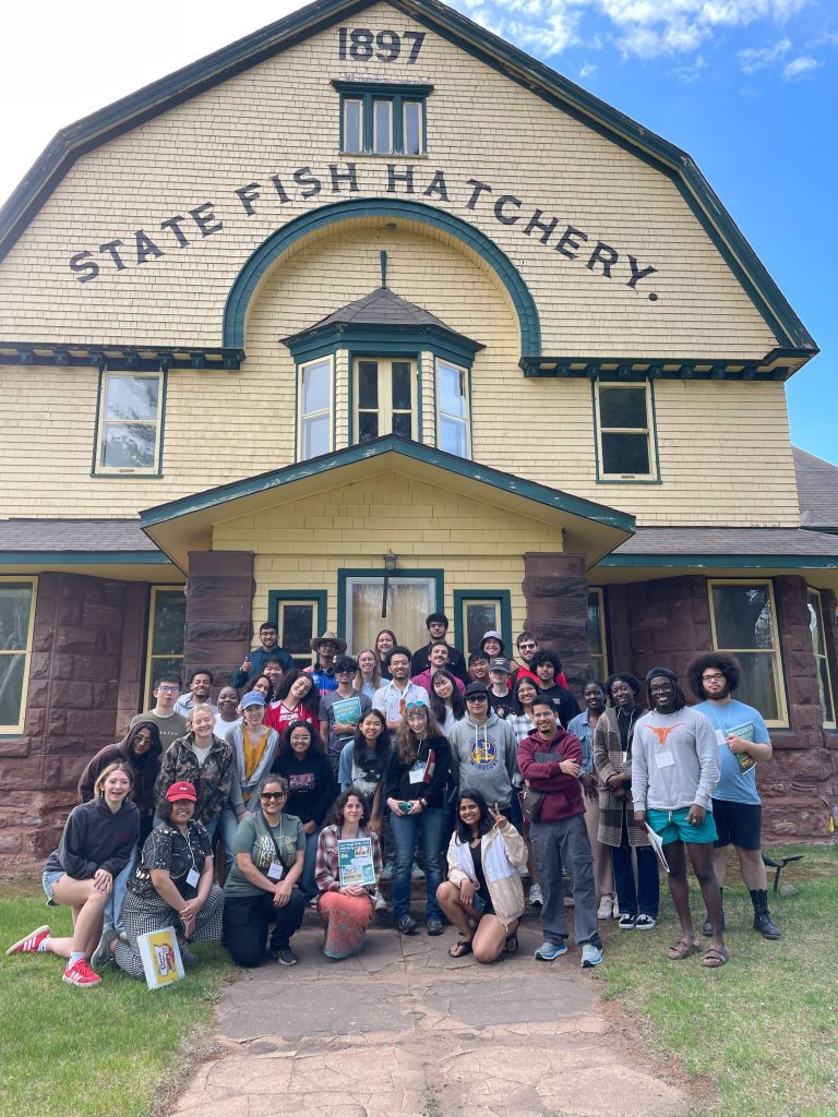 A tall, outdoor group photo of WEBT students posed in front of the original, historic Les Voigt State Fish Hatchery building.