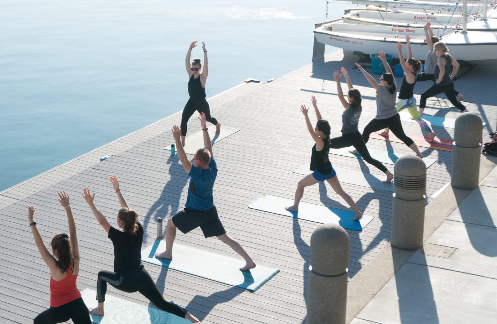 Photo of Outdoor UW Summer Yoga Class. Nine people in doing a pose with arms straight up in the air and legs in a lunged position.